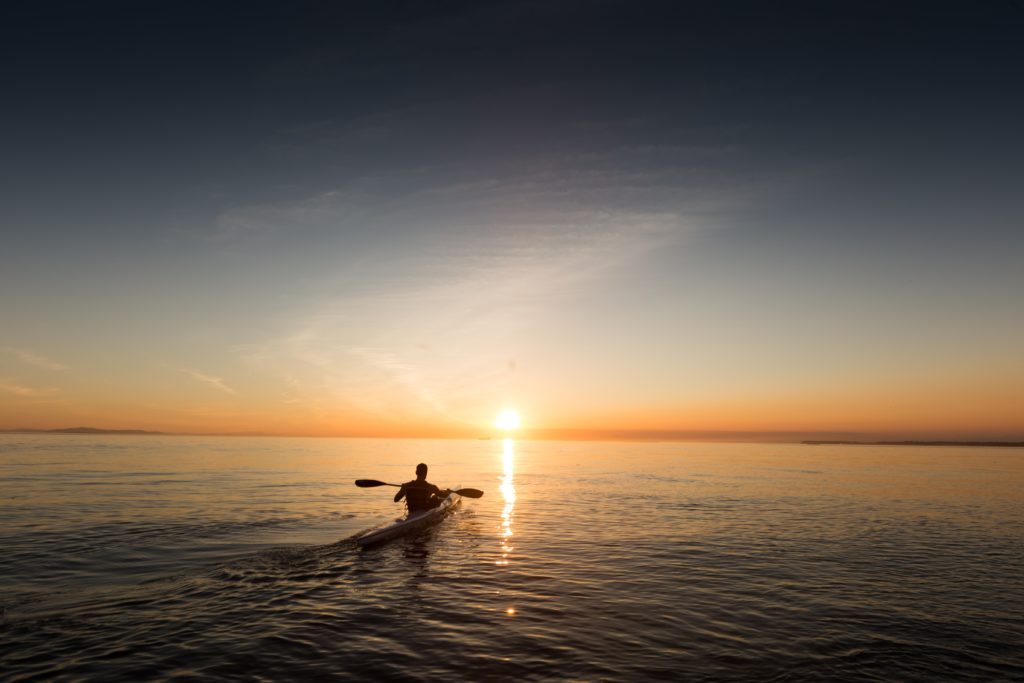 A wide open water image with a silhouette of a man paddling away from the camera in a kayak. On the horizon is the setting sun over the water.